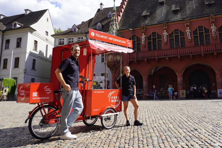 Two people stand beside a bright red tour information bike cart in a cobblestone square, with historic buildings in the background.