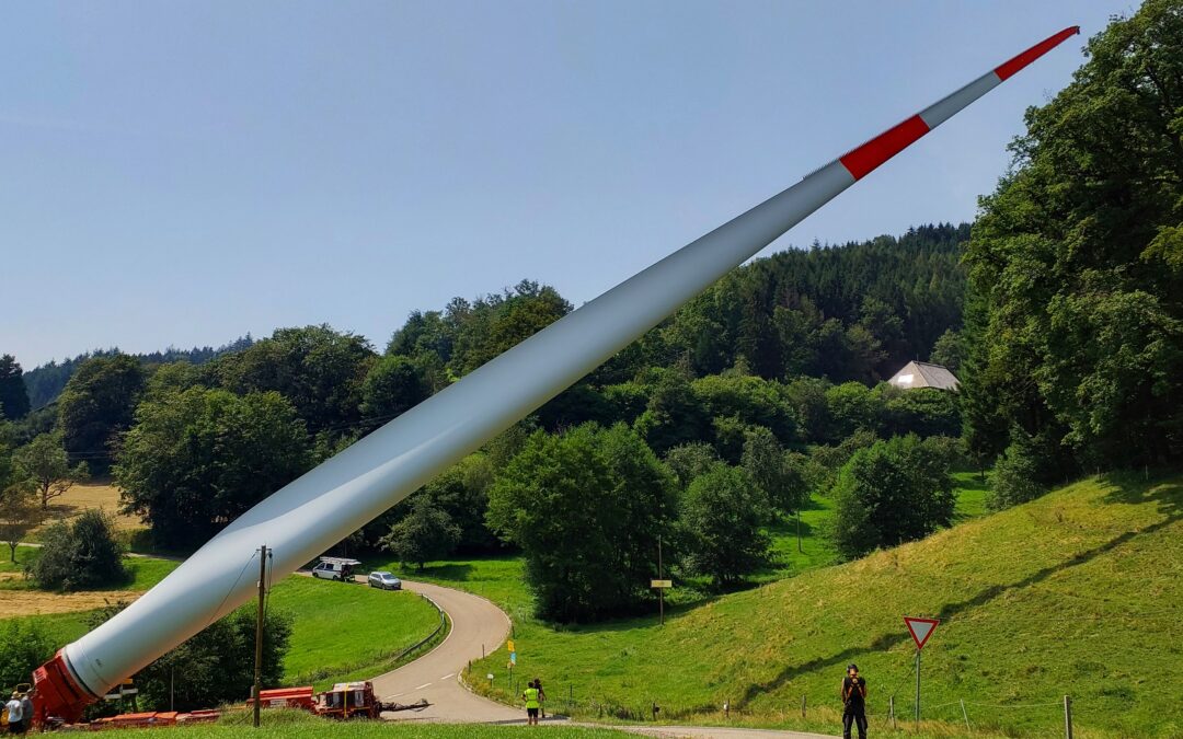 Large white wind turbine blade with red tip lying diagonally across a hillside, with workers and trucks at the base by a winding road.