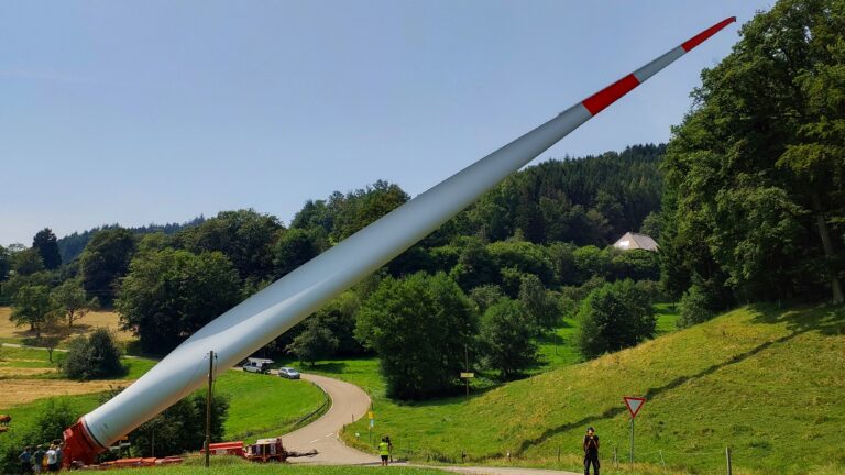 Large white wind turbine blade with red tip lying diagonally across a hillside, with workers and trucks at the base by a winding road.