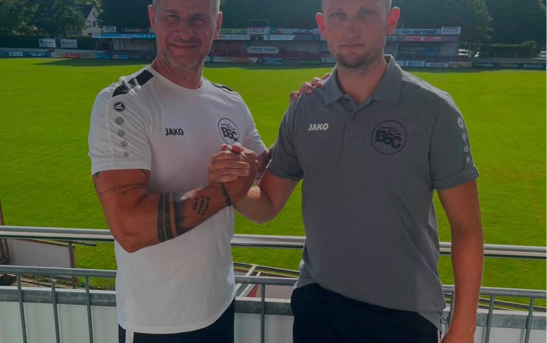 Two men in sportswear shake hands on a stadium balcony, with a green field and stands behind them on a sunny day.