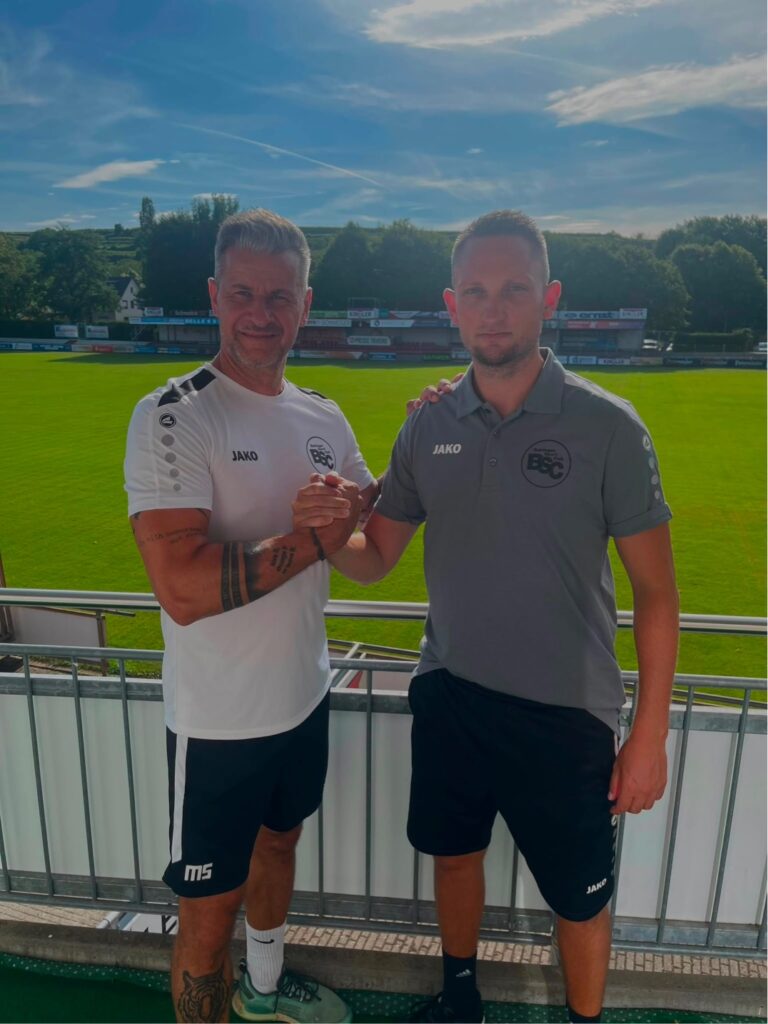 Two men in sportswear shake hands on a stadium balcony, with a green field and stands behind them on a sunny day.