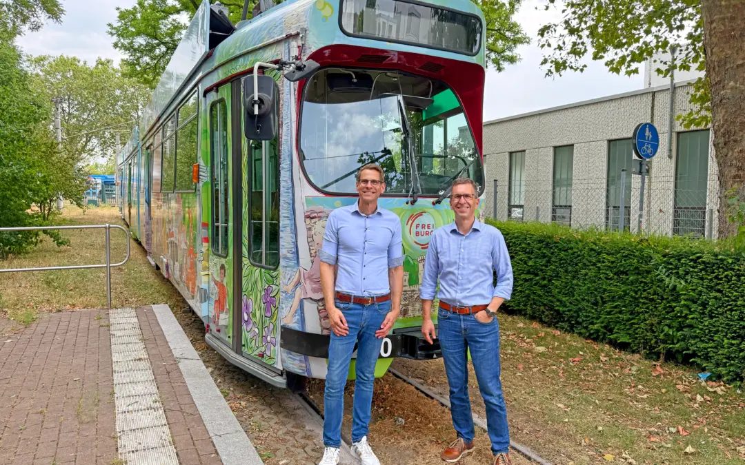 Two men in light blue shirts standing in front of a colorful tram on tracks outdoors with greenery and a modern building nearby.