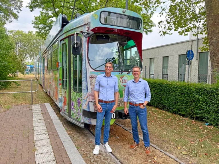 Two men in light blue shirts standing in front of a colorful tram on tracks outdoors with greenery and a modern building nearby.