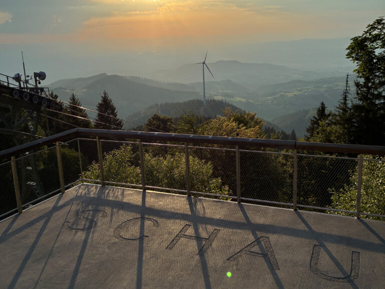 Observation deck overlooking a hazy valley with a distant wind turbine and sun rays in a golden sky.