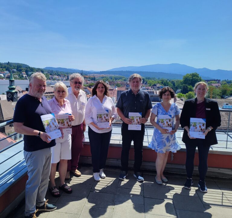 Group of adults standing on a rooftop terrace holding brochures, with a town and distant hills in the background on a sunny day.