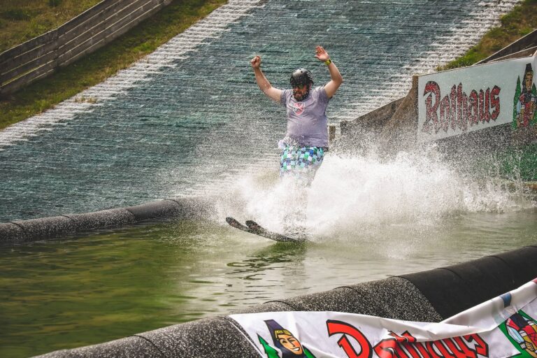 Male wakeboarder in a helmet raises his arms as he lands in a big water splash at the wakeboarding ramp.