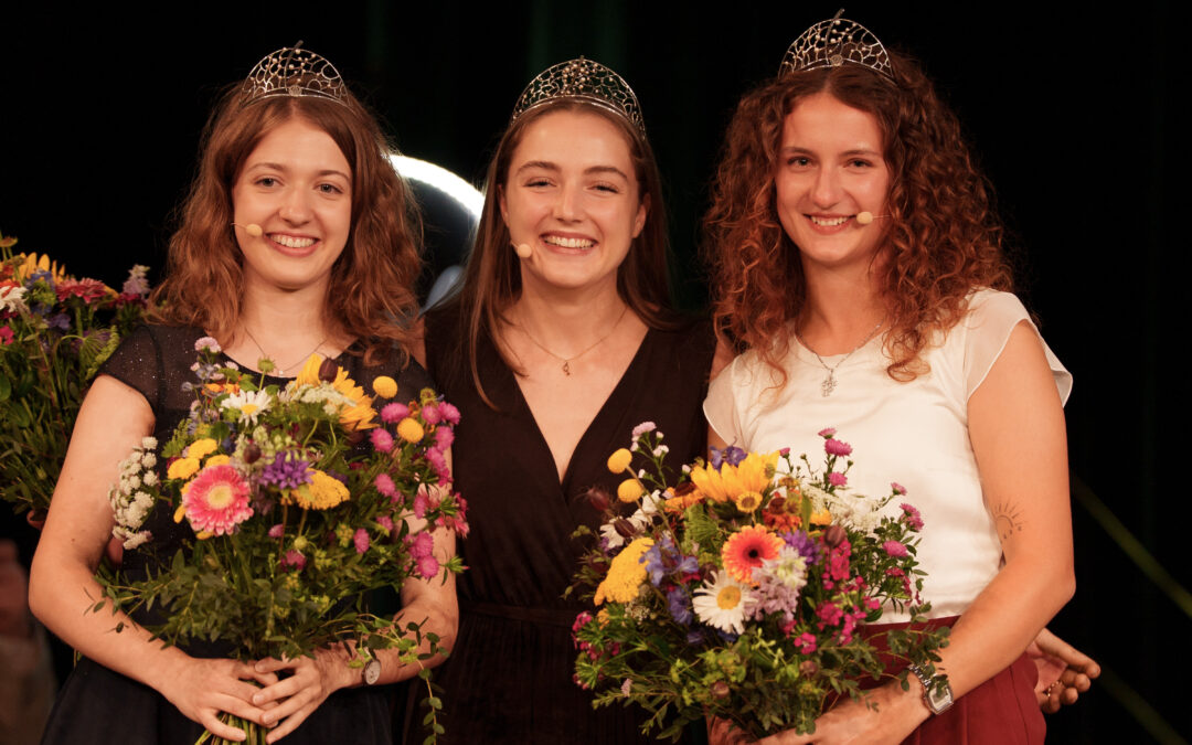 Three smiling women in evening gowns wearing tiaras, each holding colorful bouquets on stage.
