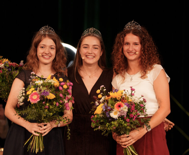 Three smiling women in evening gowns wearing tiaras, each holding colorful bouquets on stage.