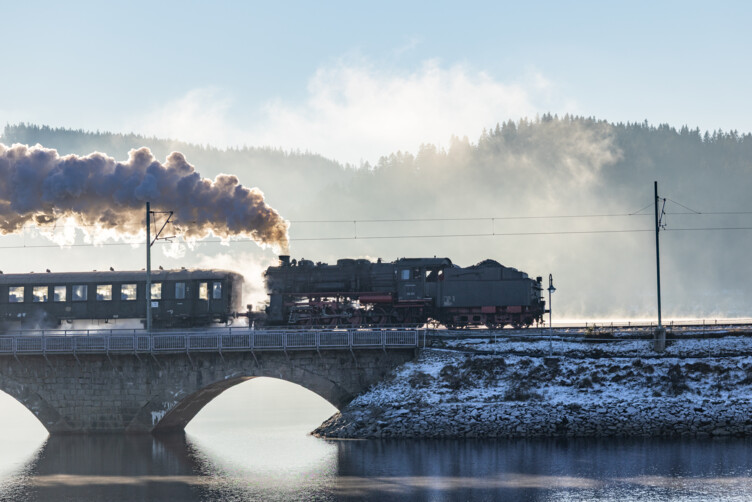 Steam locomotive pulling cars crosses a stone-arch bridge over a calm river in a snowy winter landscape smoke pluming from the engine