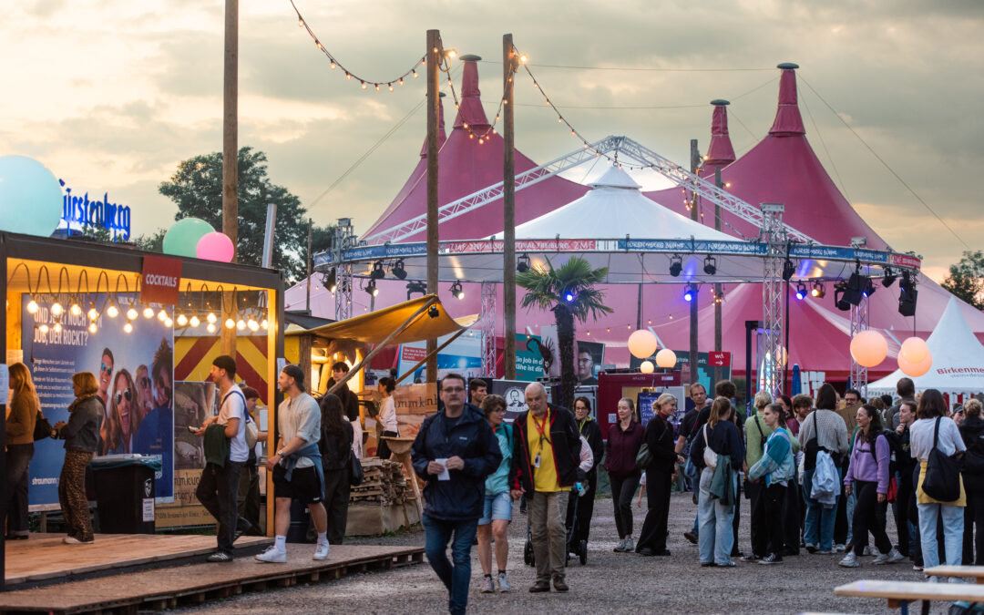 Crowd of festival-goers at an outdoor fair with pink tents, string lights, and vendor booths at dusk.