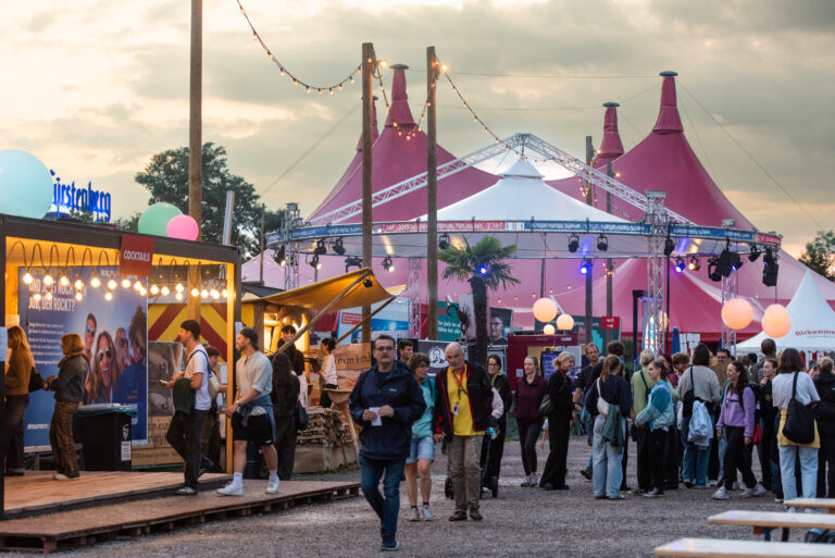 Crowd of festival-goers at an outdoor fair with pink tents, string lights, and vendor booths at dusk.