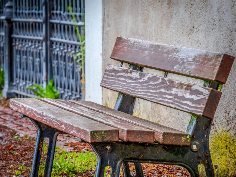 Weathered outdoor wooden bench with peeling paint and moss on the metal frame, placed beside a textured wall and iron fence.
