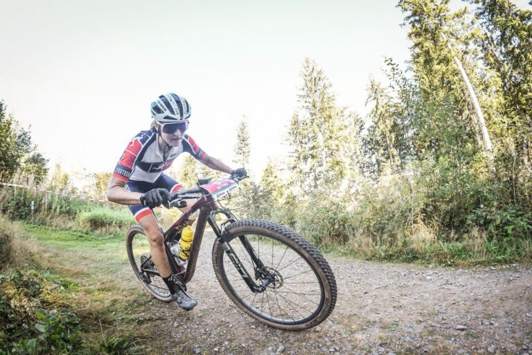 Cyclist wearing a helmet and racing jersey leans into a rough dirt trail on a full-suspension mountain bike with a yellow water bottle in the frame.