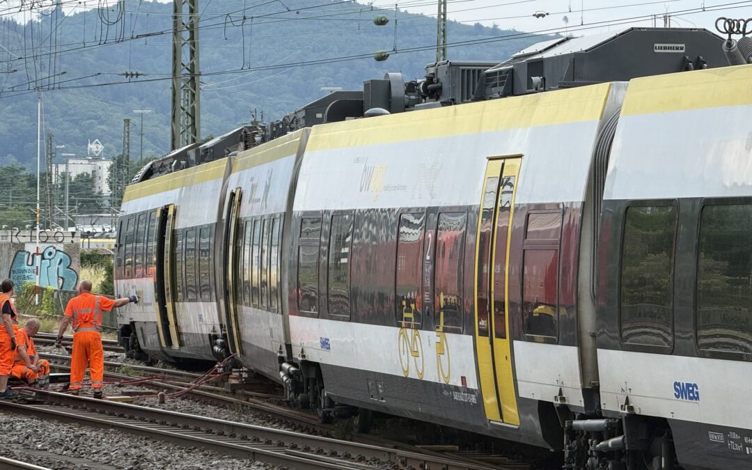 Modern passenger train on electrified tracks with overhead power lines and workers in bright orange safety vests nearby in a rail yard