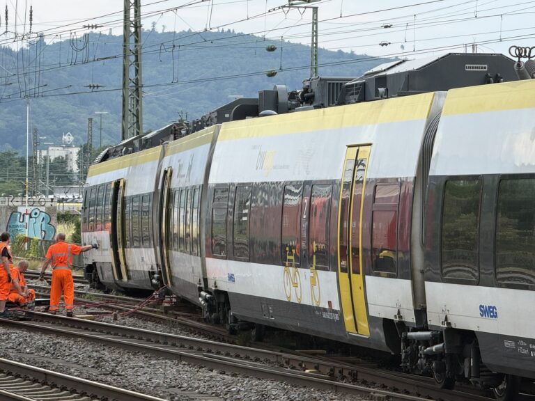 Modern passenger train on electrified tracks with overhead power lines and workers in bright orange safety vests nearby in a rail yard