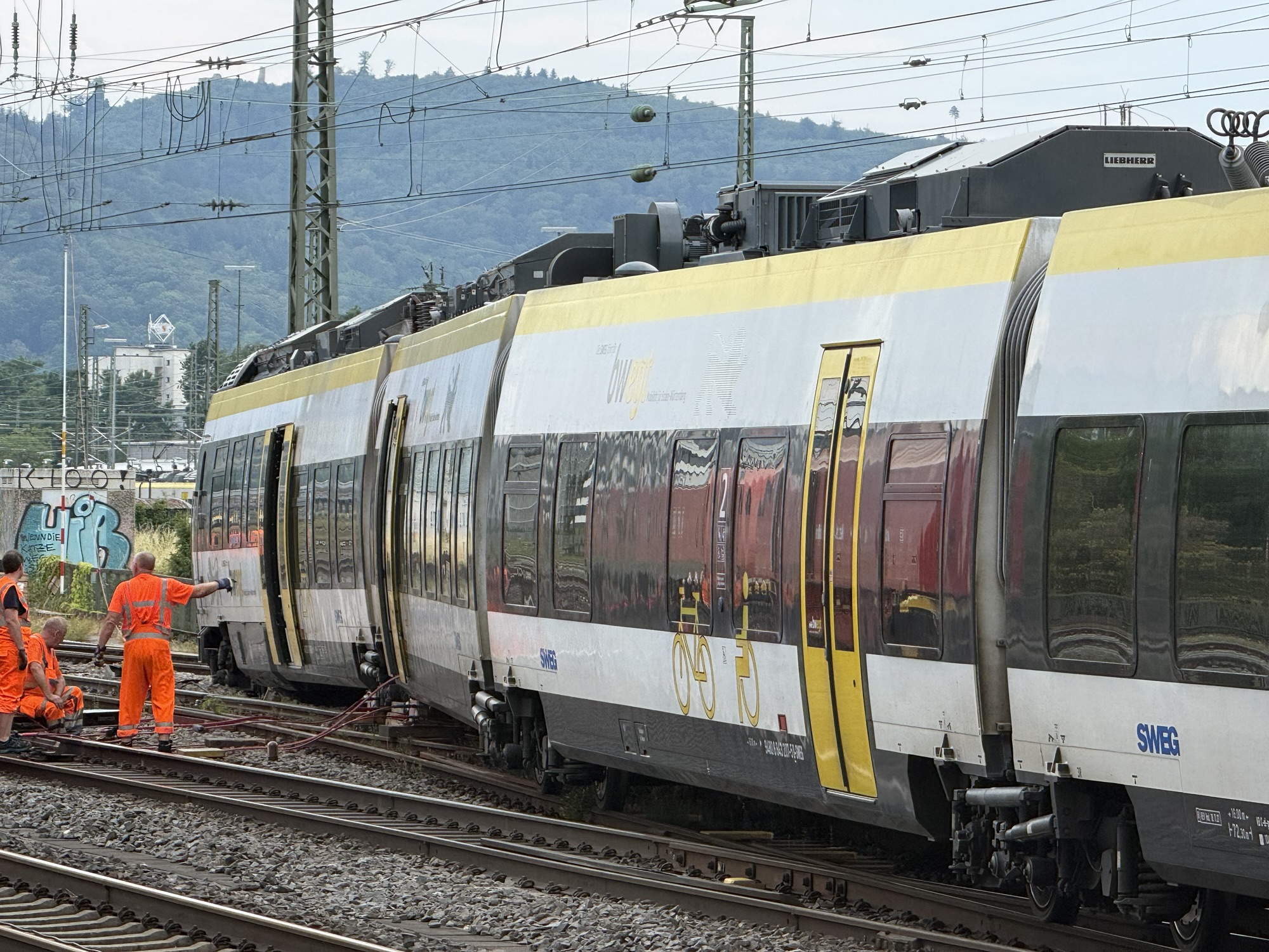 Modern passenger train on electrified tracks with overhead power lines and workers in bright orange safety vests nearby in a rail yard