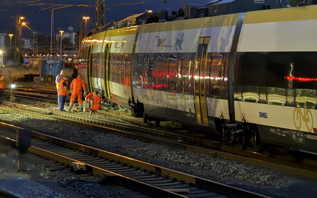 Night scene at a railway yard with a modern yellow and white train and maintenance workers in orange vests beside the tracks under overhead wires.