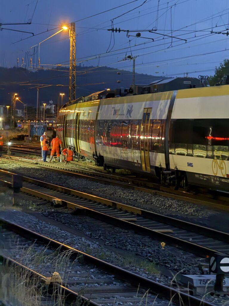 Night scene at a railway yard with a modern yellow and white train and maintenance workers in orange vests beside the tracks under overhead wires.