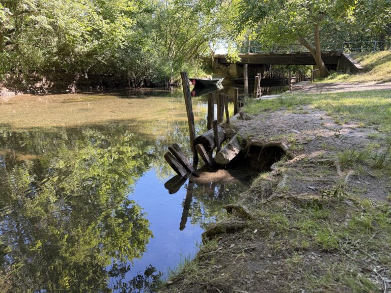 Riverside scene with calm water, reflections of trees, a weathered wooden fence along the shore, and a bridge in the background.