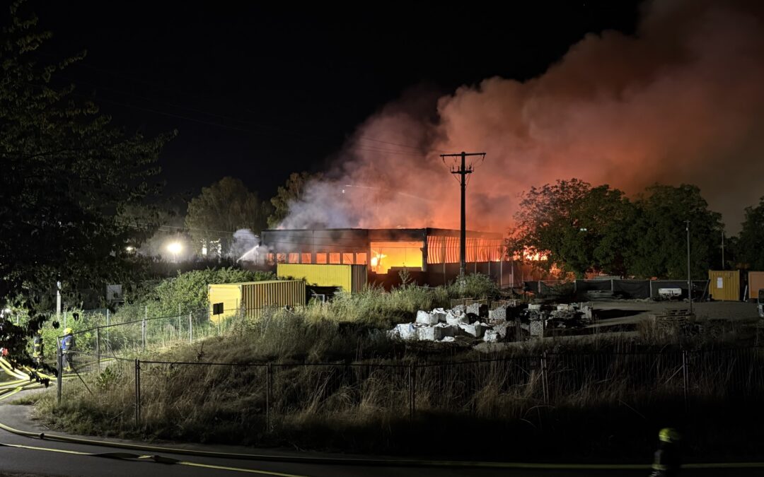 Nighttime fire engulfs an industrial building as flames and thick smoke rise; firefighters spray water from the left side.