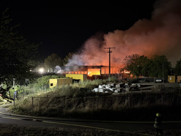 Nighttime fire engulfs an industrial building as flames and thick smoke rise; firefighters spray water from the left side.