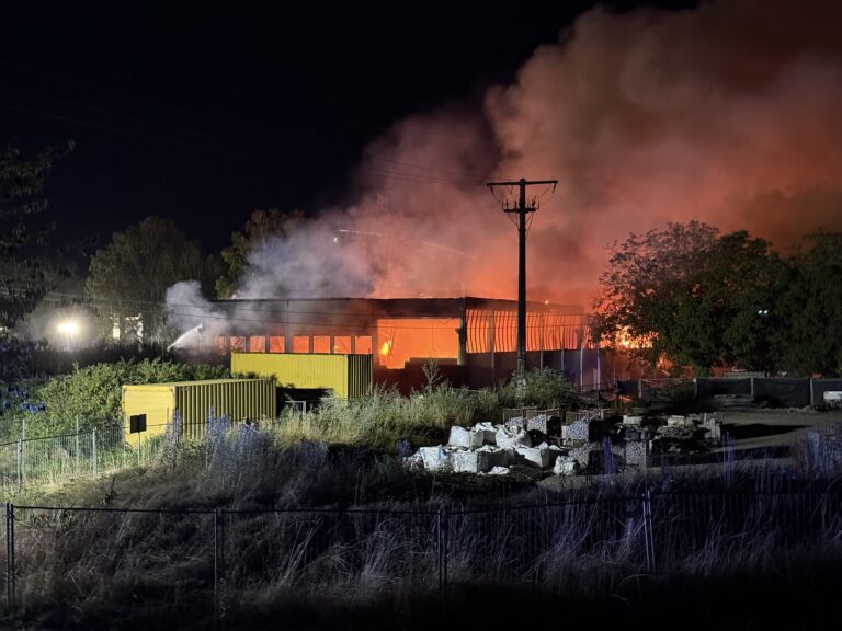 Industrial building on fire at night with orange flames and thick smoke, power pole in foreground.