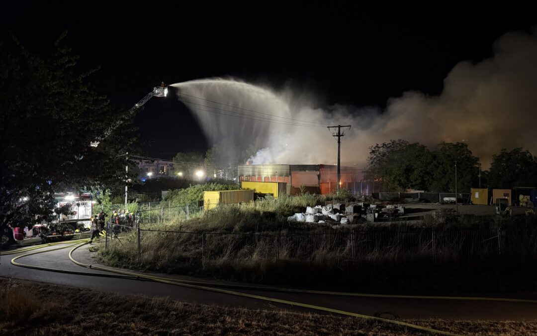 Nighttime firefighting scene: a ladder truck sprays a high arc of water onto a blaze amid thick smoke over an industrial area.