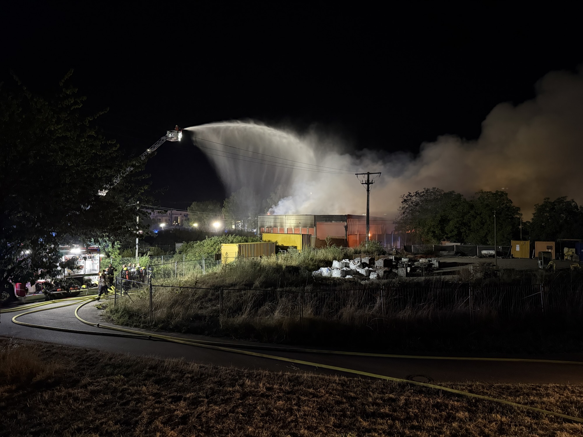 Nighttime firefighting scene: a ladder truck sprays a high arc of water onto a blaze amid thick smoke over an industrial area.