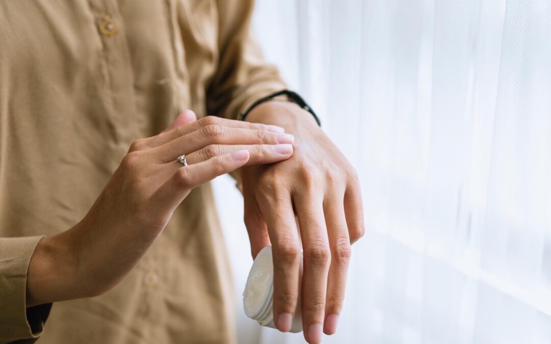 Close-up of a person applying lotion to the back of their hand from a white jar near a window