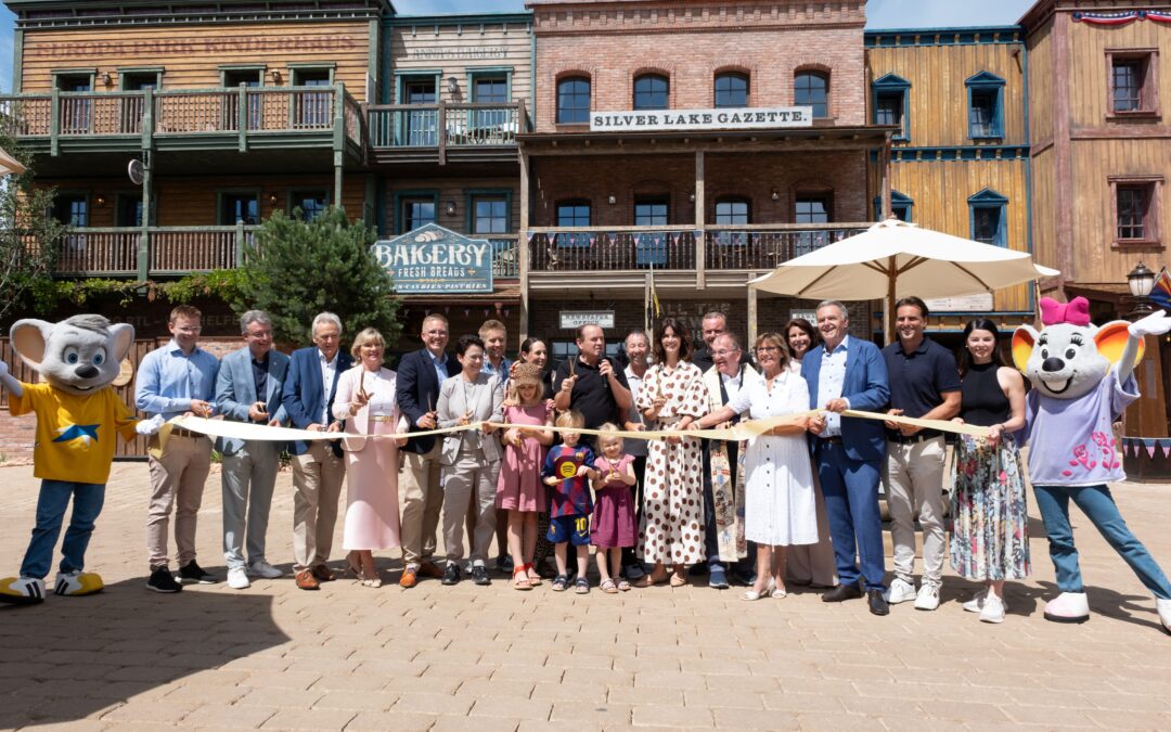 Group of people at a ribbon-cutting ceremony with mascots in a Western town setting and a long banner ribbon across the scene.