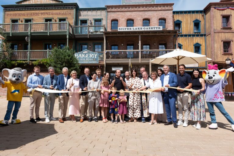 Group of people at a ribbon-cutting ceremony with mascots in a Western town setting and a long banner ribbon across the scene.