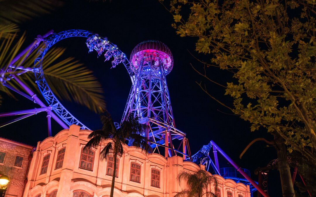 Nighttime view of a roller coaster looping above a beige building, illuminated in blue and purple lights with trees in the foreground.