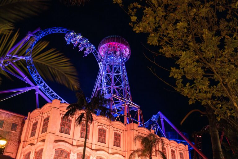 Nighttime view of a roller coaster looping above a beige building, illuminated in blue and purple lights with trees in the foreground.