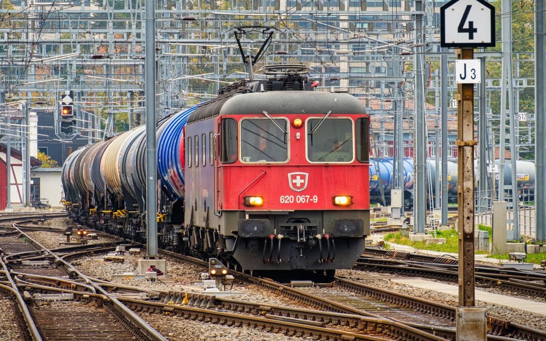Red electric locomotive leading a long freight train in a crowded rail yard with overhead wires and signals visible above the cars.
