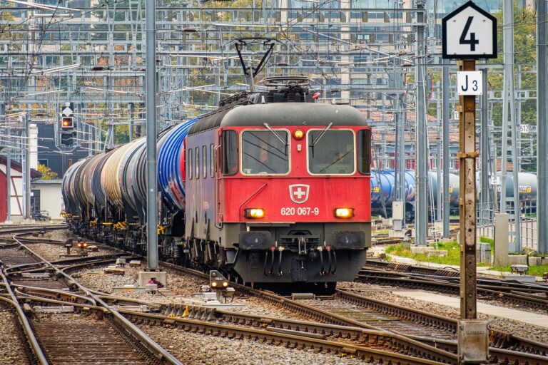 Red electric locomotive leading a long freight train in a crowded rail yard with overhead wires and signals visible above the cars.