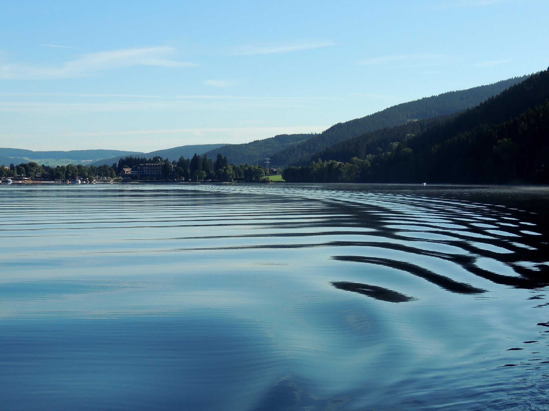 Calm lake with gentle ripples reflecting a blue sky, forested hills, and a small lakeside town on the left.