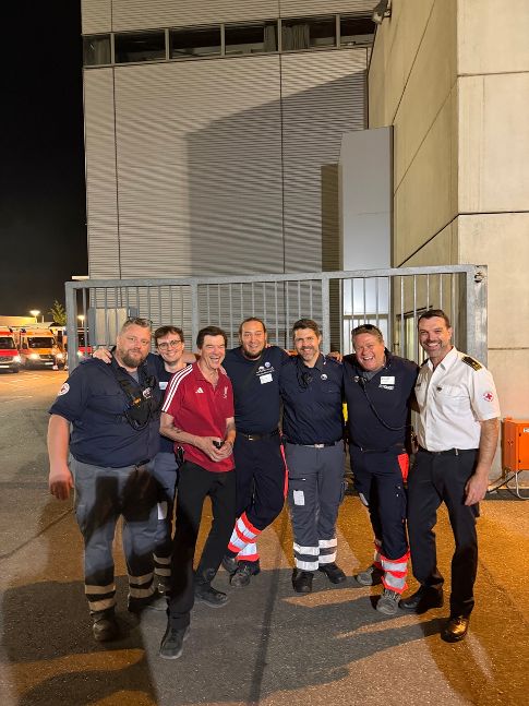 Group of eight emergency responders posing together outside a gated entrance at night, smiling and showing camaraderie in uniformed gear.