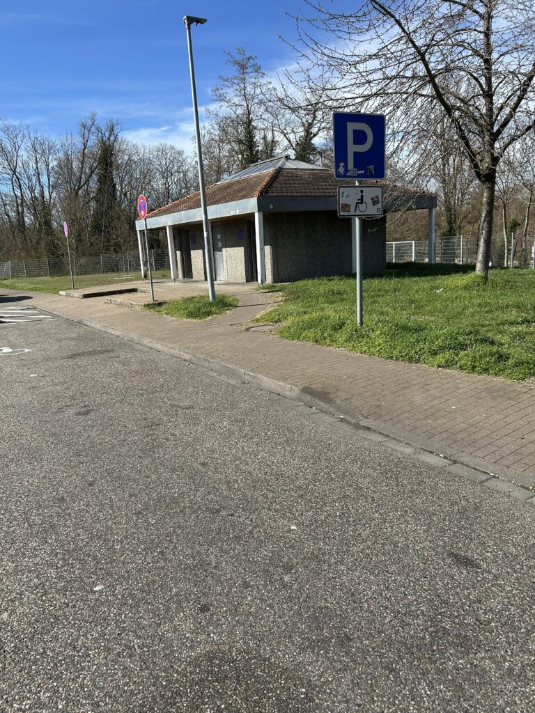 Small public restroom building with a brown tile roof beside a parking lot on a sunny day, surrounded by trees and grass.