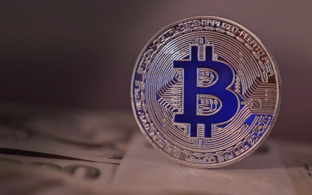 Close-up of a silver Bitcoin coin with a blue B logo, resting on a blurred surface with paper underneath.