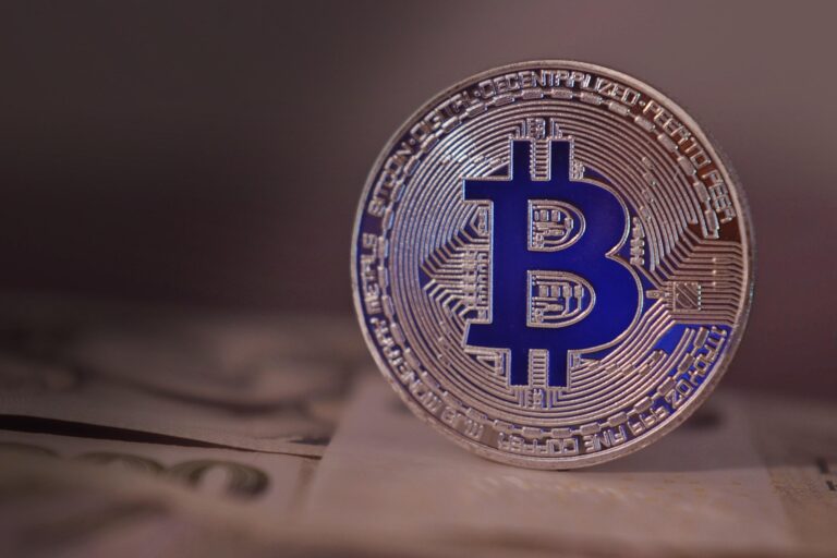Close-up of a silver Bitcoin coin with a blue B logo, resting on a blurred surface with paper underneath.