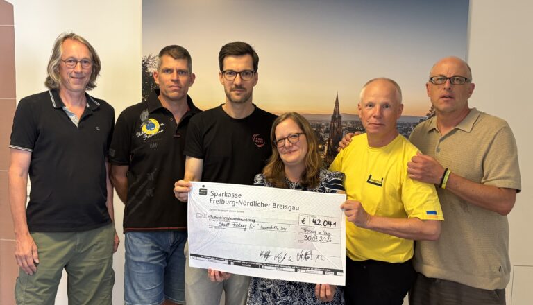 Six adults stand together indoors, holding a large donation check for €42,041 from Sparkasse Freiburg-Nördlicher Breisgau.