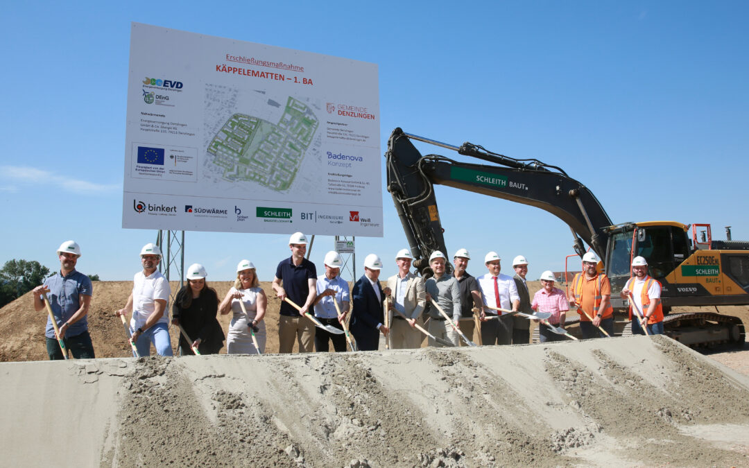 Group of construction workers and engineers with hard hats holding shovels at a site, standing in front of a large project sign and an excavator nearby