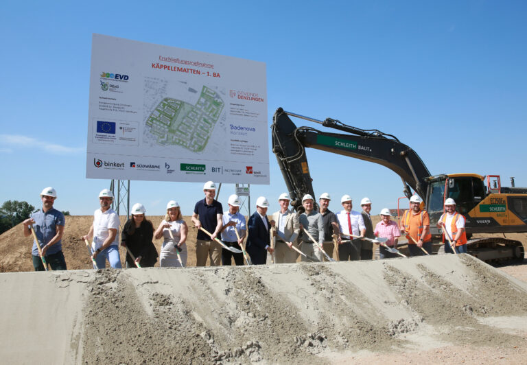 Group of construction workers and engineers with hard hats holding shovels at a site, standing in front of a large project sign and an excavator nearby