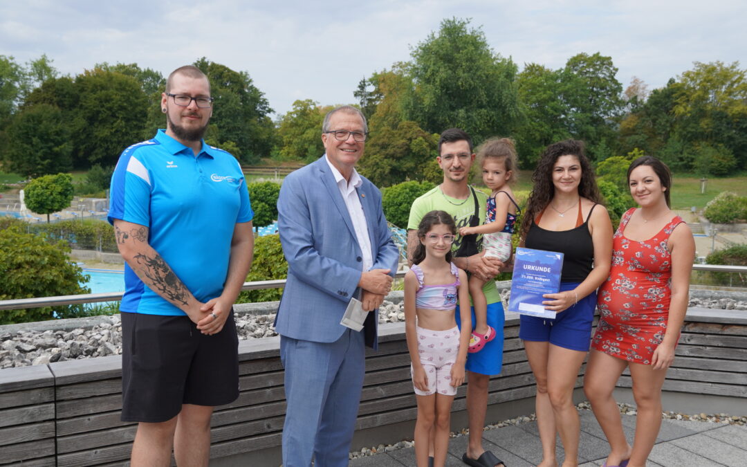 Group of seven people posing outdoors on a wooden deck, smiling at the camera, with trees in the background; a man in a blue polo, a man in a light blue suit, a man in a neon shirt holding a child, two girls, and two women (one holding a certificate).