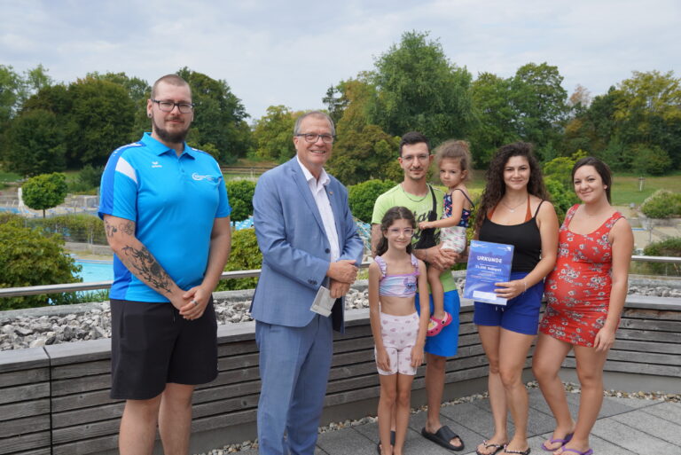 Group of seven people posing outdoors on a wooden deck, smiling at the camera, with trees in the background; a man in a blue polo, a man in a light blue suit, a man in a neon shirt holding a child, two girls, and two women (one holding a certificate).