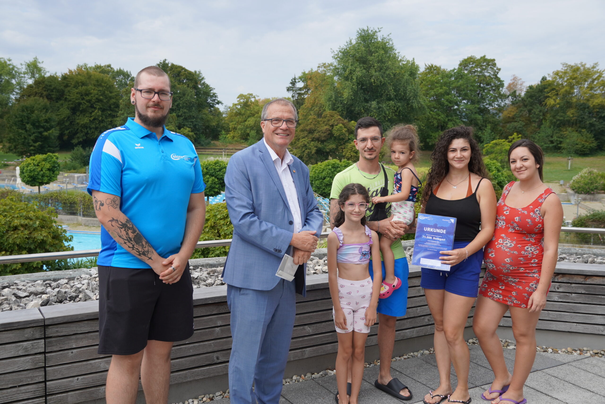 Group of seven people posing outdoors on a wooden deck, smiling at the camera, with trees in the background; a man in a blue polo, a man in a light blue suit, a man in a neon shirt holding a child, two girls, and two women (one holding a certificate).