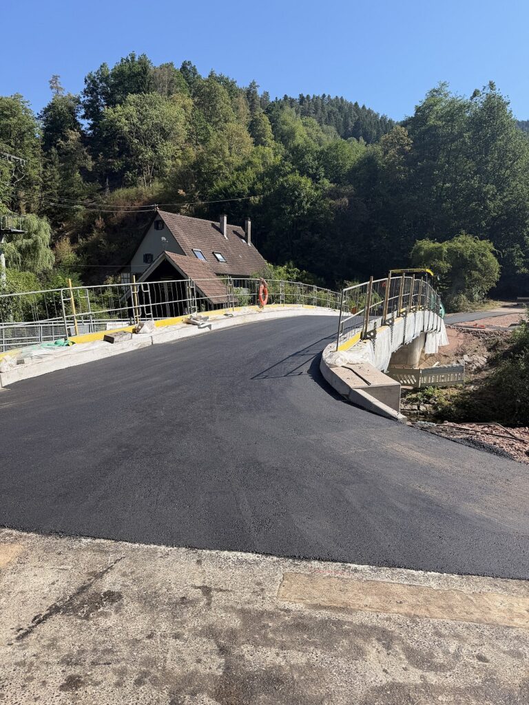 Freshly paved curved road on a hillside bridge with metal guardrails, a nearby house, and dense forest behind under a blue sky.