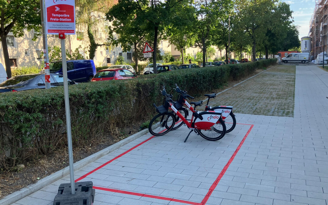 Red-and-white frelo bikes docked along a paved pedestrian path next to a trimmed hedge, with red riding lines on the ground indicating a bike lane area and a construction site in the distance.