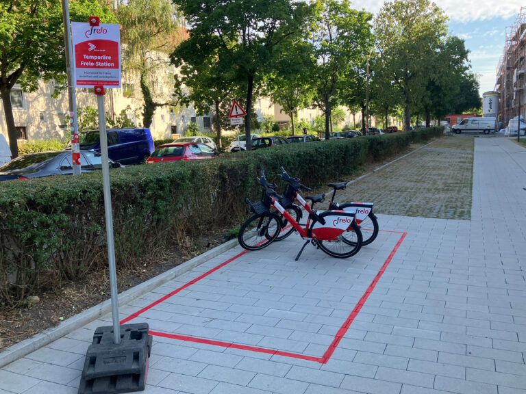Red-and-white frelo bikes docked along a paved pedestrian path next to a trimmed hedge, with red riding lines on the ground indicating a bike lane area and a construction site in the distance.