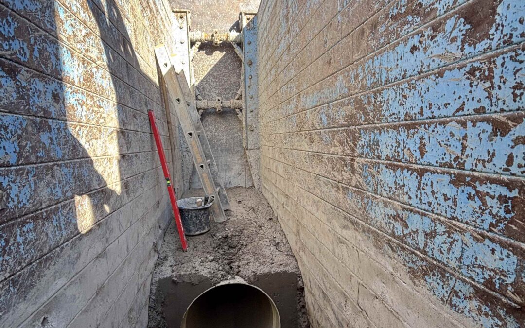Narrow construction trench between weathered concrete walls with a large cylindrical pipe resting in muddy ground; a ladder and tools are nearby, blue sky above.
